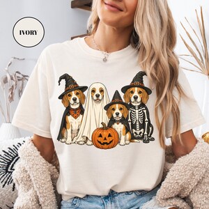 A woman wearing a white t-shirt with a Halloween-themed graphic of four dogs dressed in costumes, including a ghost, skeleton, and pumpkin.