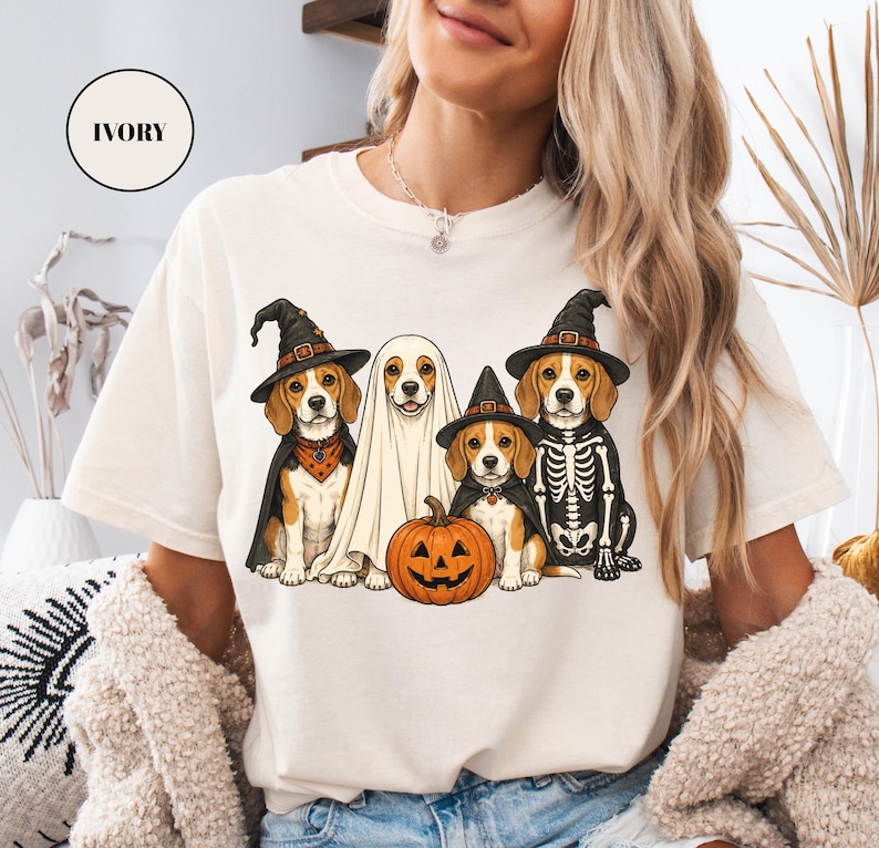 A woman wearing a white t-shirt with a Halloween-themed graphic of four dogs dressed in costumes, including a ghost, skeleton, and pumpkin.