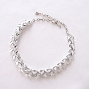 Puede incluir: Un collar de cadena de plata con un diseño trenzado. El collar está hecho de metal y tiene un cierre de broche.