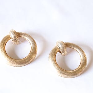 May include: A pair of gold-toned hoop earrings with a textured finish. The earrings have a unique design with a small loop at the top.