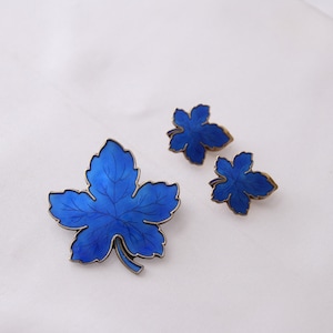 Puede incluir: Un conjunto de pendientes de hoja de esmalte azul y un broche a juego. Las hojas están detalladas con un contorno plateado y tienen un centro azul oscuro.