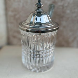 May include: A clear glass sugar bowl with a silver-colored lid and rim. The glass has a cut pattern, and the lid is topped with a decorative finial. A small spoon rests on the side.