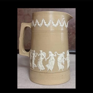 May include: Tan ceramic pitcher with a handle and spout. The pitcher is decorated with white relief figures of people dancing and a white garland design around the top. The base has a textured pattern.