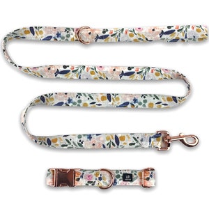 May include: A white dog collar and leash set with a floral pattern in shades of pink, blue, and yellow. The hardware is rose gold.