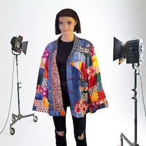 May include: A young person wearing a blue denim jacket with colorful patchwork fabric. The jacket has a patch that says "One of a Kind Heart". The person is standing in front of a white background with two studio lights.