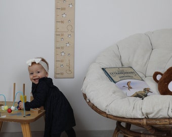 May include: A wooden growth chart with a white background and a cutout star design. The chart is marked in centimeters and features the name "Gustave" written in cursive. A toddler in a black dress is standing next to the chart.