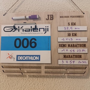 Peut inclure: Un tableau en bois avec un dossard de course à pied et une liste de records personnels pour différentes distances. Le dossard est bleu avec le numéro 006 et le texte "Kalenji find your rhythm enjoy your run" et "By Decathlon". Les records sont pour 5 km, 10 km, semi-marathon et marathon. Les temps sont écrits en minutes et en secondes.