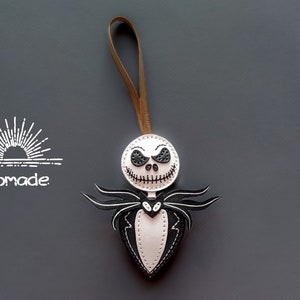 May include: White leather ornament with a black stitched Jack Skellington face and a black stitched bow tie. The ornament is attached to a brown leather cord.