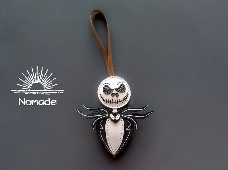 May include: White leather ornament with a black stitched Jack Skellington face and a black stitched bow tie. The ornament is attached to a brown leather cord.