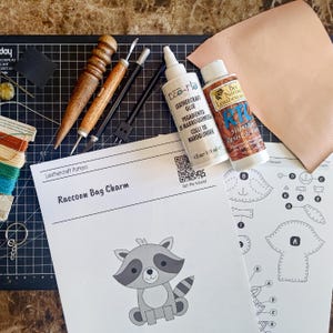 May include: A leathercraft kit for a raccoon bag charm, including a printed pattern with a QR code, a piece of light tan leather, a metal key ring, colorful thread, a wooden burnisher, a metal awl, a craft knife, and bottles of glue and finish.