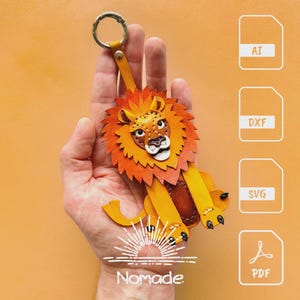 May include: A handcrafted leather lion keychain in shades of orange and yellow. The lion has a detailed mane, face, and paws. The keychain includes a metal ring and a leather strap. The word "Nomade" is printed on the bottom.