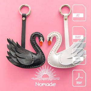 May include: Two leather swan keychains, one black and one white, with orange beaks, hang from silver rings. The keychains are on a pink background. The word "Nomade" is at the bottom.