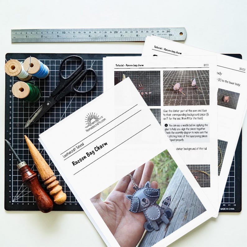 May include: A leathercraft tutorial for a raccoon bag charm, featuring printed instruction sheets with diagrams, a metal ruler measuring 30 cm, black scissors, two wooden burnishers, and two spools of thread on a black cutting mat with grid lines.