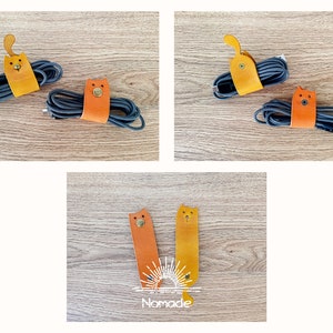 May include: Three leather cat-shaped cable organisers in orange and yellow. The organisers are on a wooden surface. The text "Nomade" is visible in the bottom centre of the image.