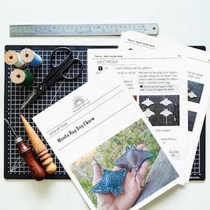May include: A crafting setup featuring tools and instructions for leather manta ray bag charms. Includes a ruler, scissors, spools of thread, leather tools, and printed tutorial sheets. The tutorial is from Nomadecrafts.com.
