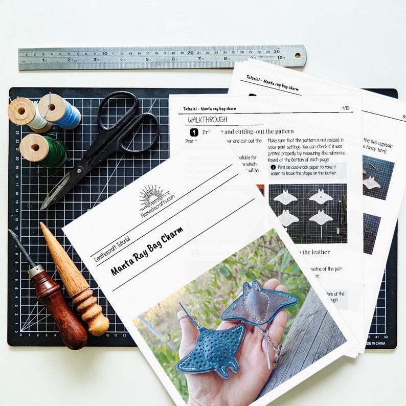 May include: A crafting setup featuring tools and instructions for leather manta ray bag charms. Includes a ruler, scissors, spools of thread, leather tools, and printed tutorial sheets. The tutorial is from Nomadecrafts.com.