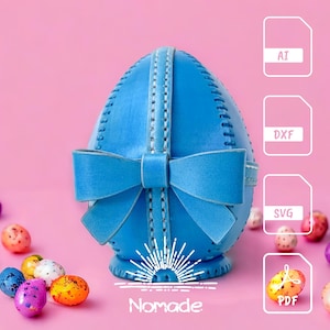 Könnte beinhalten: Ein blaues Leder-Osterei mit Schleife, verziert mit Nähten. Das Ei liegt auf einer rosa Oberfläche mit weißem Hintergrund. Der Text "Nomade" ist unter dem Ei sichtbar. Das Bild enthält auch eine Reihe von Symbolen für verschiedene Dateiformate: AI, DXF, SVG und PDF.