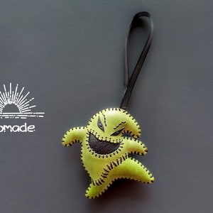 May include: A green felt ornament shaped like a character from the movie Nightmare Before Christmas. The character has a stitched-on black mouth and eyes. The ornament is hanging from a black leather cord.