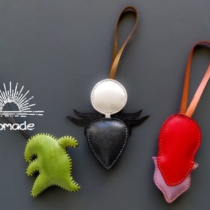 May include: Three leather ornaments with different designs. The first is a green ghost, the second is a black heart with a white circle and wings, and the third is a red heart with a purple flower.