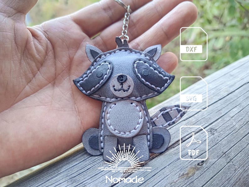 May include: A gray and dark gray leather raccoon keychain featuring stitched details, a round belly patch, and a striped tail. The keychain includes a metal ring and chain. The image displays icons for DXF, SVG, and PDF file formats and the text Nomade.