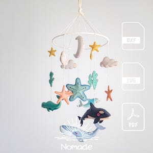 Peut inclure: Un mobile suspendu composé de créatures marines en feutrine, dont une baleine, des étoiles de mer et du corail, ainsi qu'une lune et des nuages suspendus à un cadre circulaire blanc. L'image inclut des icônes DXF, SVG, PDF et le texte Nomade.