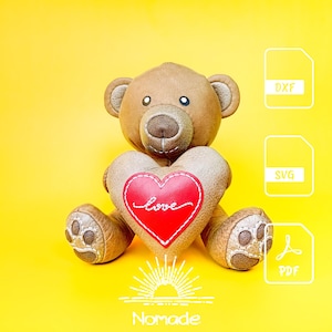 May include: A brown teddy bear holding a heart-shaped pillow with a red center and the word love embroidered in white. The bear has stitched paw pads and dark eyes. To the right are three white file icons labeled DXF, SVG, and PDF.