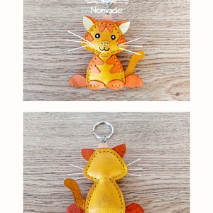 May include: A yellow leather cat keyring with orange accents. The cat is sitting with its tail curled around its legs. The keyring has a silver ring and the word "Nomade" is stitched on the back.