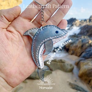 Puede incluir: Un charm de bolso de cuero con forma de delfín con un llavero plateado. El delfín es gris y blanco con ojos azules y costuras blancas. El texto "Leathercraft Pattern" y "Dolphin Bag Charm" son visibles.