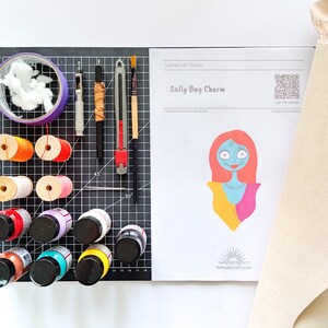 May include: A leathercraft pattern for a Sally Bag Charm from Nightmare Before Christmas. The pattern includes a color illustration of Sally, a QR code, and the text "Get the Tutorial". The image also shows a variety of craft supplies, including leather, paint, thread, and tools.
