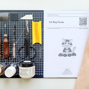 May include: A leathercraft pattern for a cat bag charm, featuring a black and white illustration of a cat. The pattern is on a white sheet of paper with the title "Cat Bag Charm" and a QR code. The image also shows a selection of leatherworking tools, including a leather awl, a stitching awl, a hole punch, a pair of scissors, a spool of thread, and a bottle of leather glue.