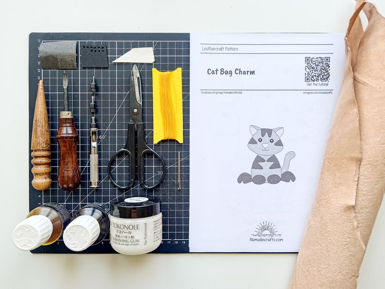 May include: A leathercraft pattern for a cat bag charm, featuring a black and white illustration of a cat. The pattern is on a white sheet of paper with the title "Cat Bag Charm" and a QR code. The image also shows a selection of leatherworking tools, including a leather awl, a stitching awl, a hole punch, a pair of scissors, a spool of thread, and a bottle of leather glue.