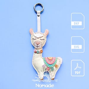 Könnte beinhalten: Ein cremefarbener Lama-Schlüsselanhänger mit genähten Details, einem bunten Sattel mit rosa, türkisfarbenen und orangefarbenen Akzenten sowie einem Metallring. Das Design enthält kleine Knöpfe am Hals und den Text DXF, SVG, PDF und Nomade.