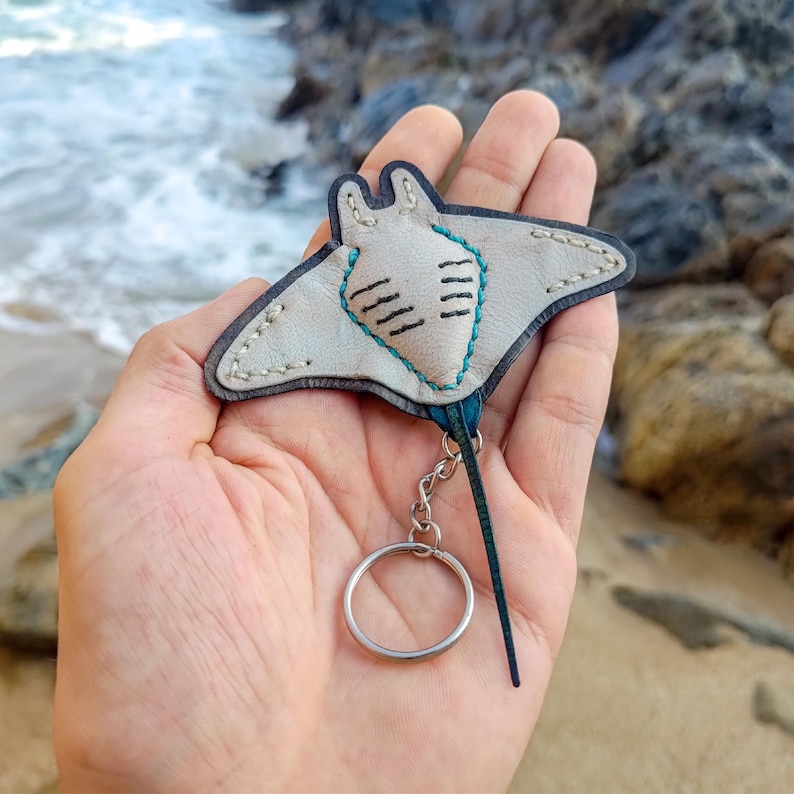 May include: A handmade leather keychain featuring a stingray design. The stingray is light grey with dark blue trim and teal stitching. The keychain has a silver-coloured ring and a dark blue tail.