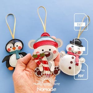 Puede incluir: Tres adornos colgantes con forma de pingüino, un oso con un bastón de caramelo y un muñeco de nieve, con detalles cosidos, botones y lazos dorados. La imagen incluye iconos de formato DXF, SVG y PDF, además del texto Nomade.