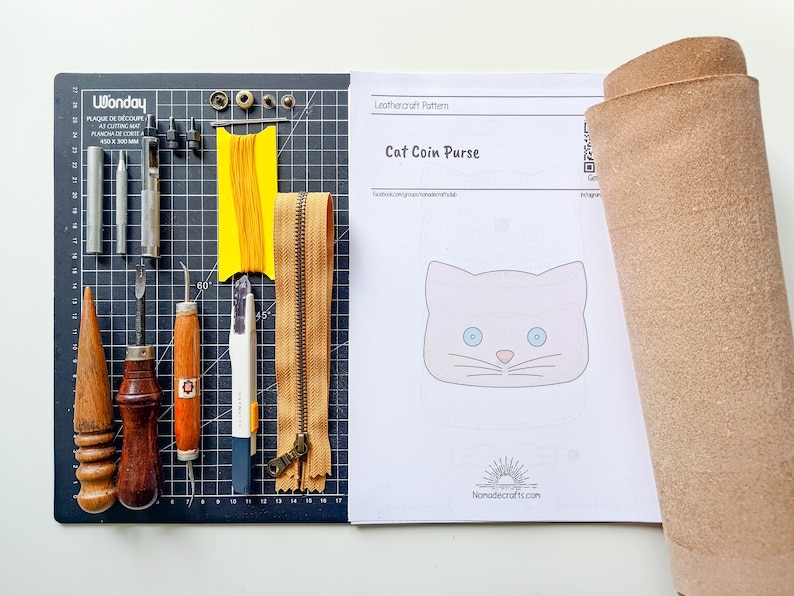 May include: A leathercraft pattern for a cat coin purse, with a template, tools, and a piece of leather. The tools include a leather punch, awl, stitching tools, a ruler, a cutting mat, and a zipper.