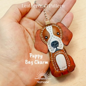 Puede incluir: Un charm de bolso de cachorro de cuero marrón y blanco con un llavero plateado. El charm tiene un diseño cosido y el texto "Puppy Bag Charm" y "Leathercraft Pattern". El charm se sostiene en una mano.