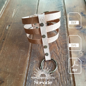 May include: A light tan, multi-layered strap accessory featuring an embossed paisley pattern, secured by four dark metal rivets and a circular ring at the base. The image includes file icons labelled DXF, SVG, and PDF, along with the text Nomade.