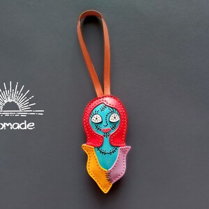 May include: A colorful leather keychain featuring a character with red hair, blue skin, and yellow and pink clothing. The character has stitched details and white eyes. The keychain has a brown leather strap.