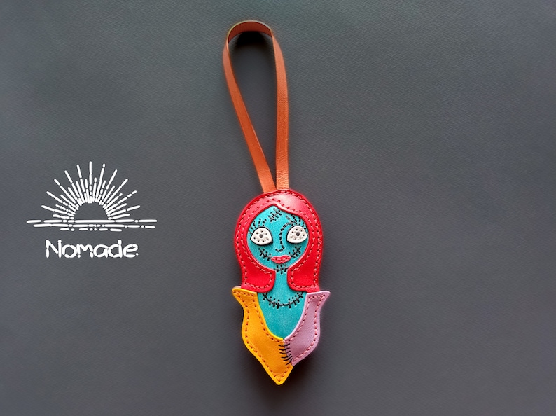 May include: A colorful leather keychain featuring a character with red hair, blue skin, and yellow and pink clothing. The character has stitched details and white eyes. The keychain has a brown leather strap.