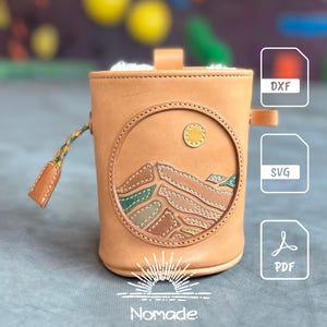 Può includere: Una borsa a secchiello in pelle color cuoio con un design circolare in rilievo raffigurante un paesaggio con un sole giallo e cime montuose verdi e marroni. La borsa presenta una linguetta intrecciata e icone di file DXF, SVG e PDF.