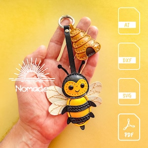 Op de afbeelding: Een leren bijensleutelhanger met een geel en zwart lichaam, witte vleugels en een lachend gezicht. De sleutelhanger bevat een kleine bijenkorfbedel en een zwarte riem. Het woord "Nomade" is zichtbaar.