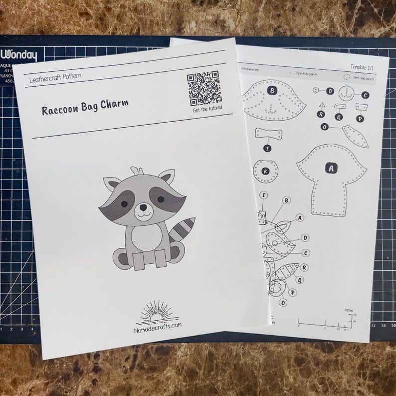 May include: A white paper leathercraft pattern for a Raccoon Bag Charm featuring a cartoon raccoon illustration, a QR code, and the text Nomadecrafts.com. A second sheet shows labeled pattern pieces with a visible 2-inch scale ruler at the bottom.
