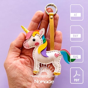 Op de afbeelding: Een kleurrijke leren eenhoorn sleutelhanger met een goudkleurige metalen ring en een gele leren riem. De eenhoorn heeft een witte body, een roze hart en een regenboog manen en staart. Het woord "Nomade" staat onderaan.