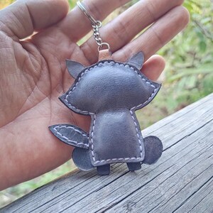 May include: A gray leather keychain shaped like a stylized animal with pointed ears and a bushy tail. The piece features visible light-colored stitching along the edges and is attached to a silver-toned metal split ring by a small chain.