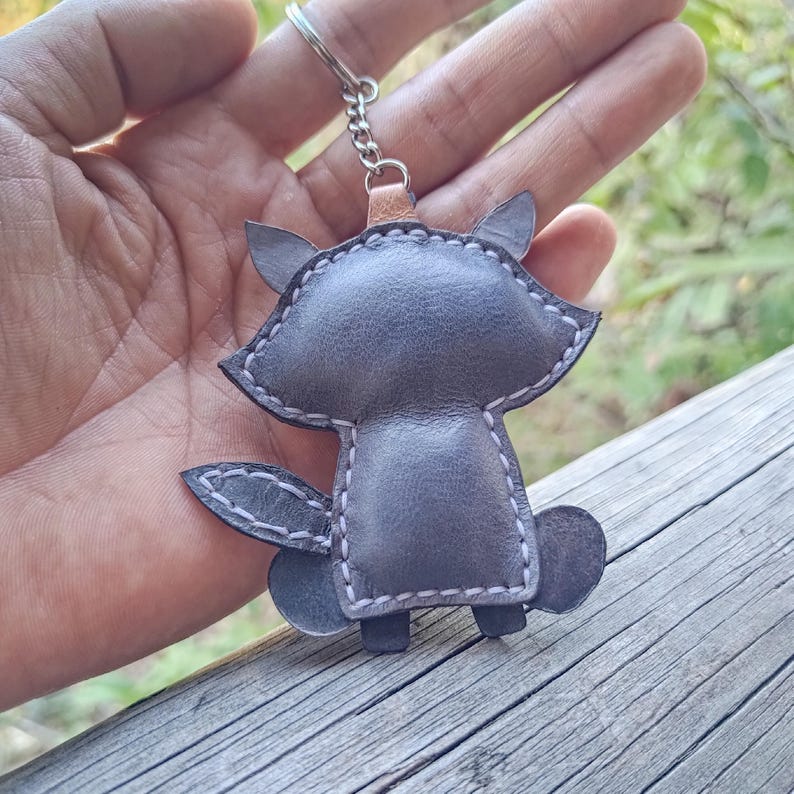 May include: A gray leather keychain shaped like a stylized animal with pointed ears and a bushy tail. The piece features visible light-colored stitching along the edges and is attached to a silver-toned metal split ring by a small chain.