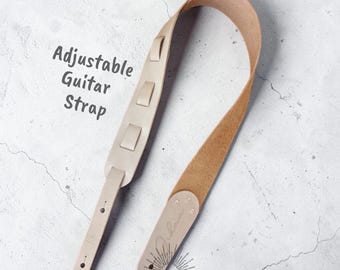 May include: A light beige adjustable guitar strap made of leather. The strap has a padded shoulder section with three rectangular accents. The text "Leathercraft Pattern" and "Adjustable Guitar Strap" are visible. The strap is branded with the name "Nomade".