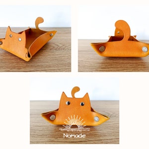 May include: A small, orange leather tray shaped like a cat. The tray has a cat face with two black eyes and a small tail. The tray has a white sun design on the bottom and the word "Nomade" printed below it.
