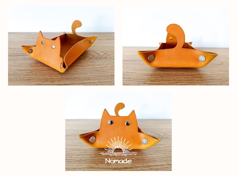 May include: A small, orange leather tray shaped like a cat. The tray has a cat face with two black eyes and a small tail. The tray has a white sun design on the bottom and the word "Nomade" printed below it.