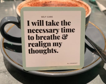 Könnte beinhalten: Eine hellgrüne Karte mit dem Text "SELF CARE" und "ALIGNMENT" oben und unten auf der Karte. Der Text "I will take the necessary time to breathe & realign my thoughts." ist in schwarzer Fettschrift auf der Karte. Die Karte liegt auf einer grauen Untertasse mit goldenem Rand. Im Hintergrund steht eine Tasse Kaffee.