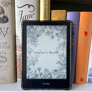 May include: A black Kindle e-reader with a floral design on the screen, displaying the text "meghan's kindle." The e-reader is in a protective case and is surrounded by a collection of books on a shelf.